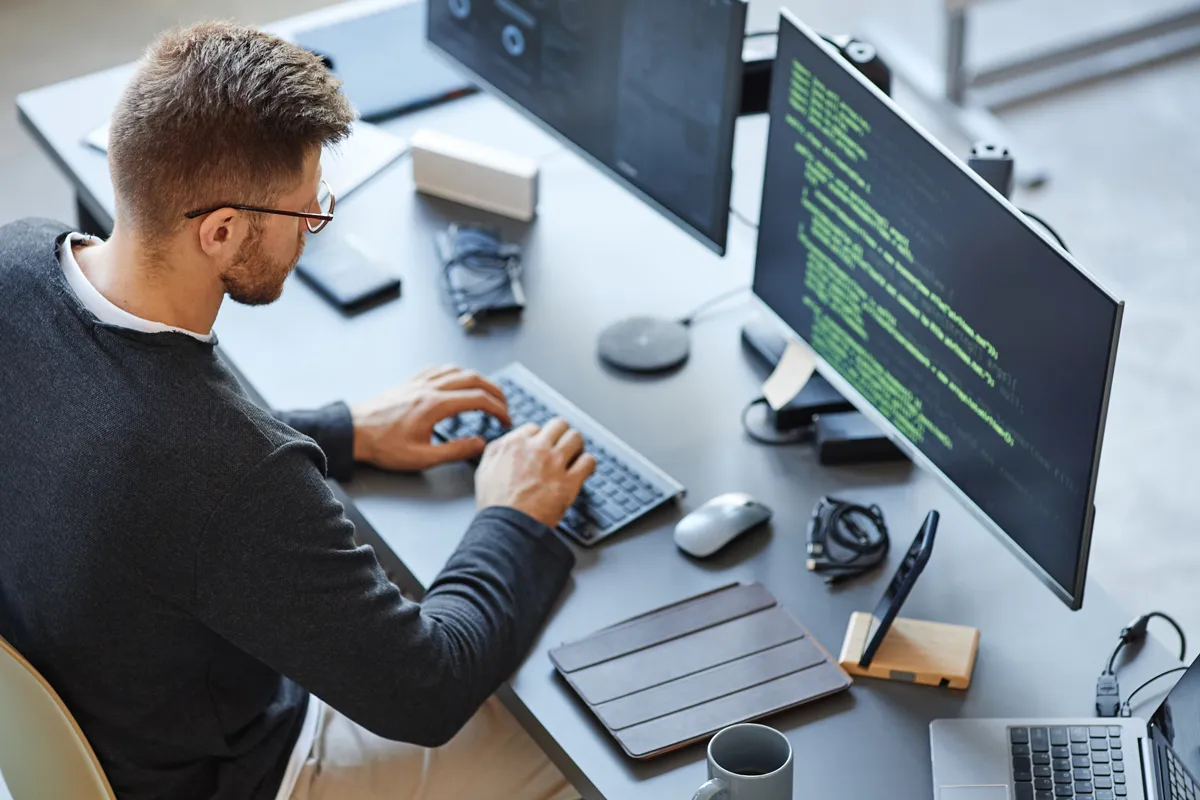 Starte bei uns als Fachinformatiker/-in bei uns und spezialisiere Dich in einer Fachrichtung Deiner Wahl. Foto eines jungen Mannes, der vor 2 Monitoren sitzt und an einem Quellcode arbeitet.